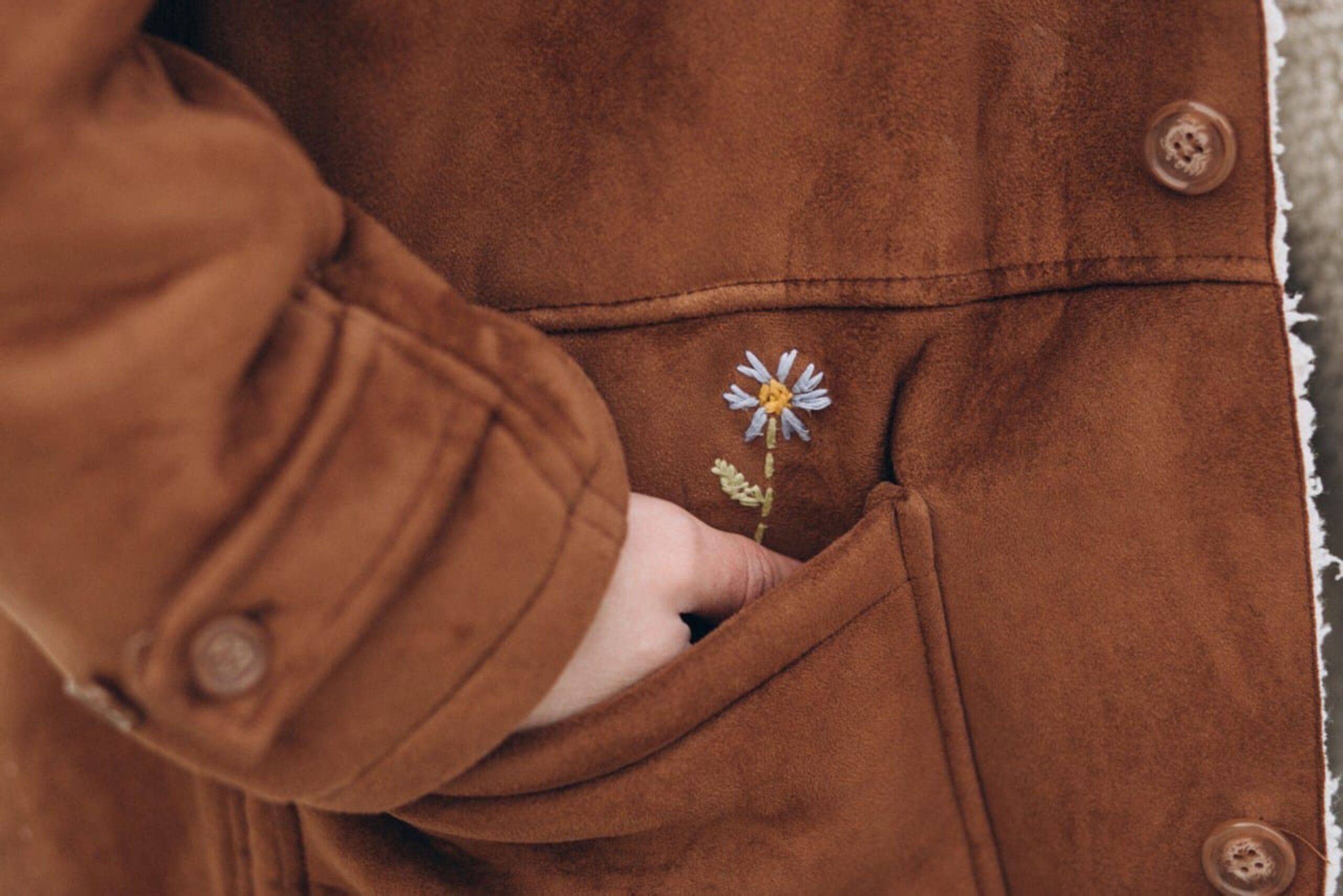 A close photo of a woman's suede jacket pocket, where her hand is being placed, revealing a small embriodered flower.