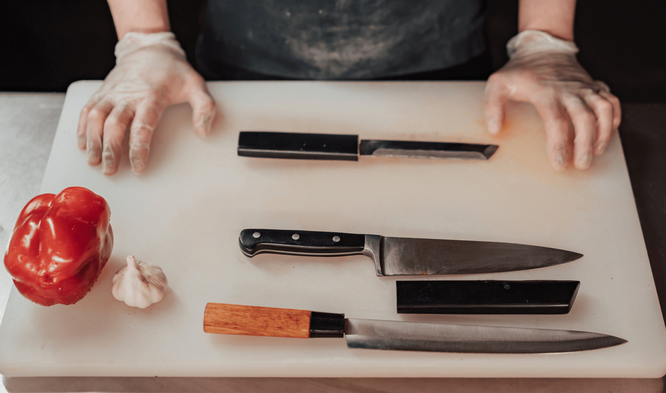 An above shot of a person's gloved hands on top ofa  cutting board, four chef's knives nearby.